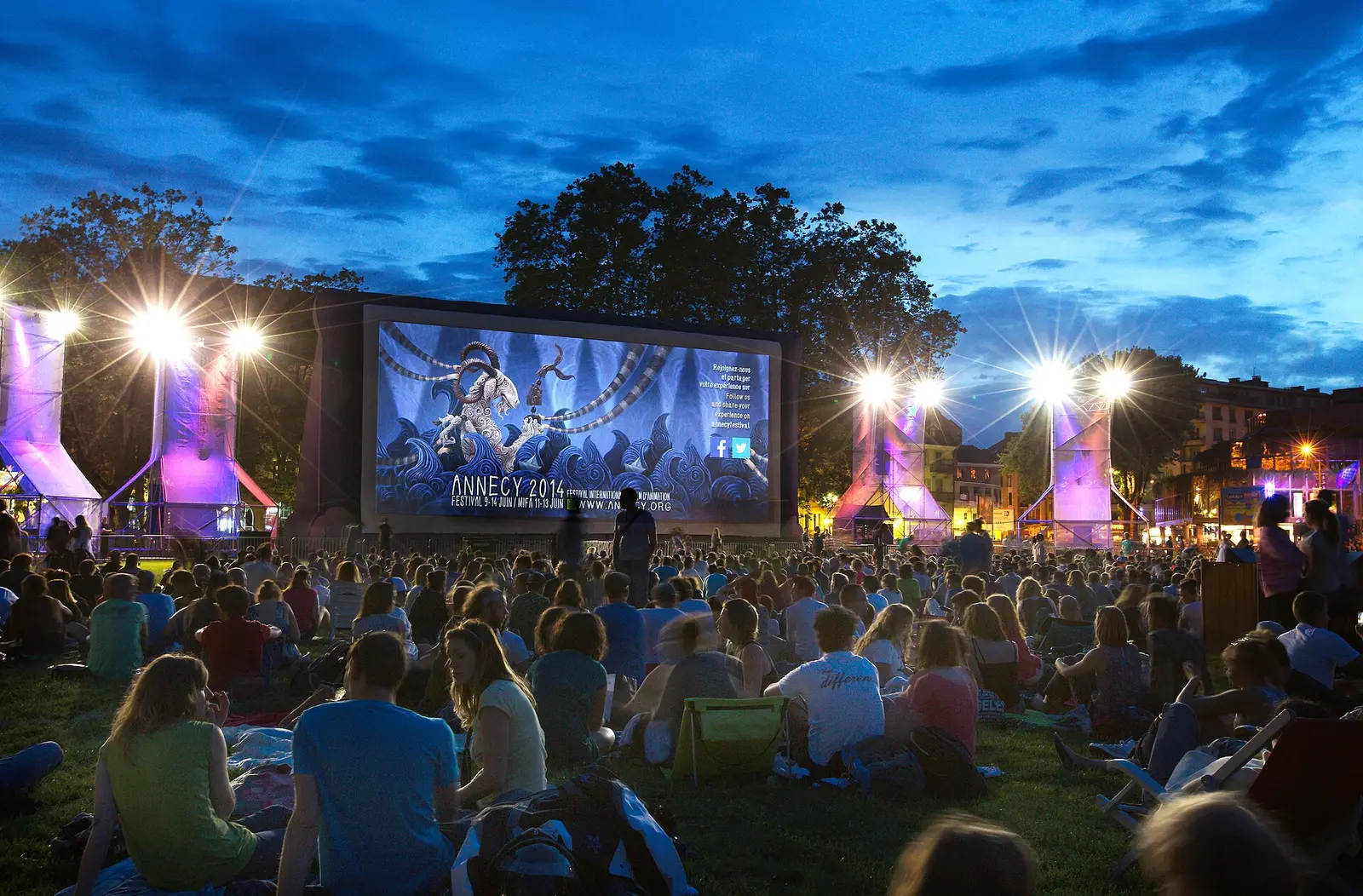 Audience on the grass watching an open-air film screening at dusk beside the lake in Annecy