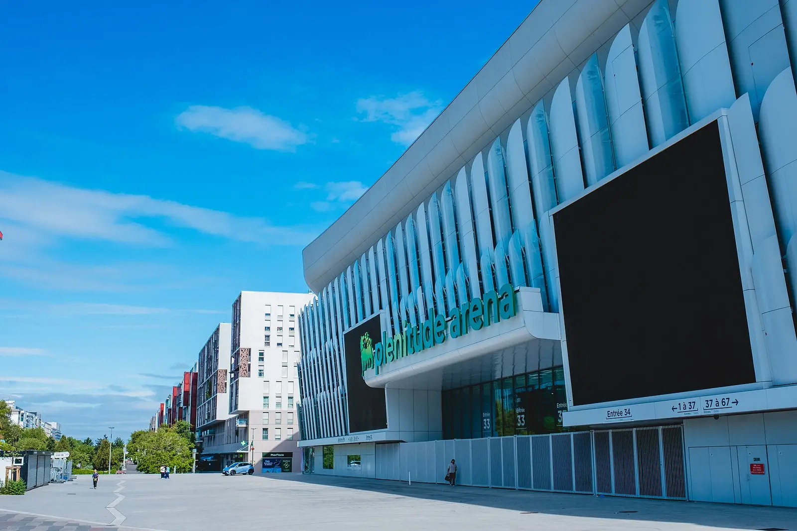 Curved white facade of the Plenitude Arena in Nanterre with a large screen and blue sky